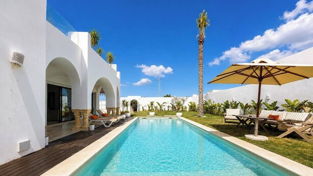 A swimming pool with lounge chairs and umbrellas, surrounded by white walls and palm trees. - Villa Hanine S3 Piscine (Hammamet)