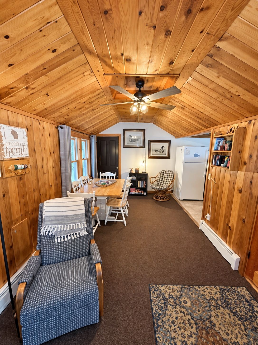 A cozy room with a wooden ceiling, a dining table with chairs, a wicker chair, a refrigerator, and a bookshelf.