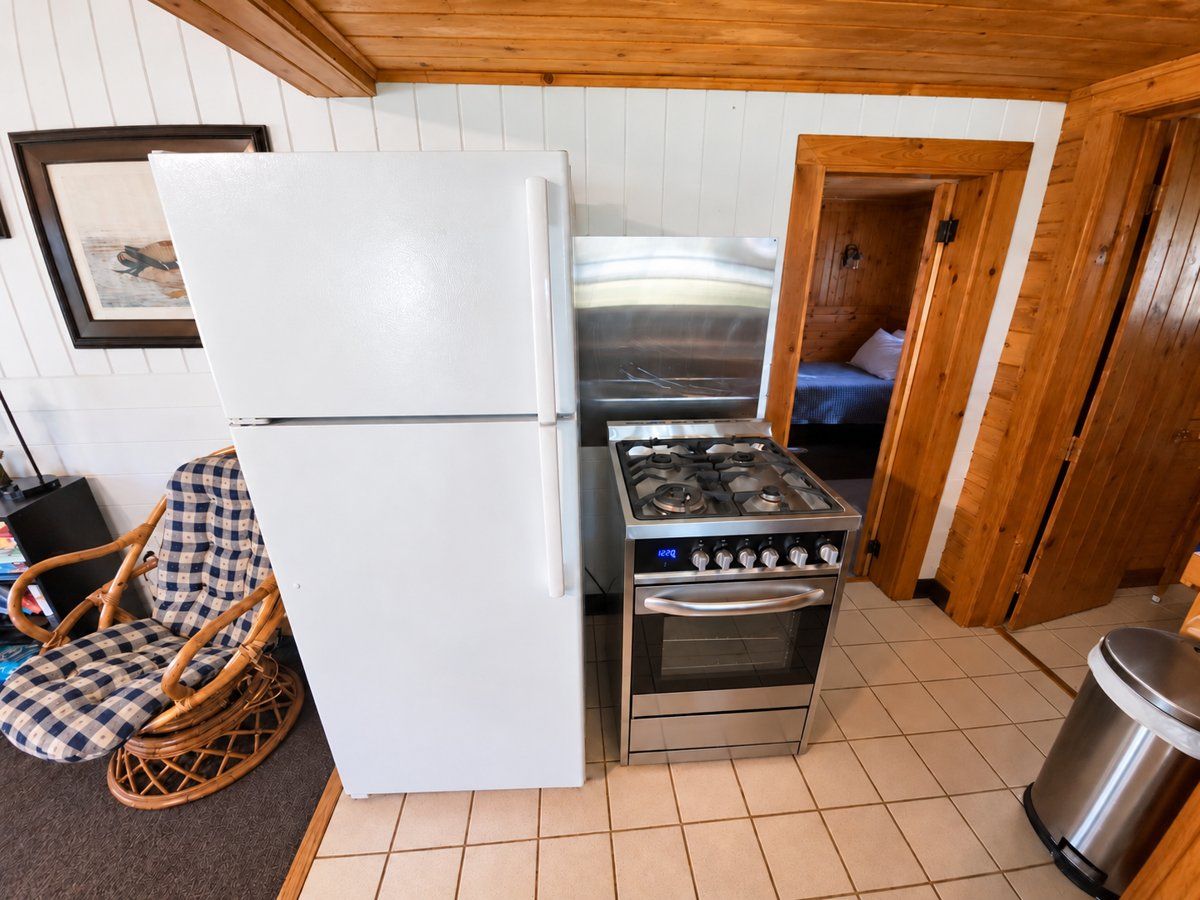 A compact kitchen with a refrigerator, stove, and a wicker chair.
