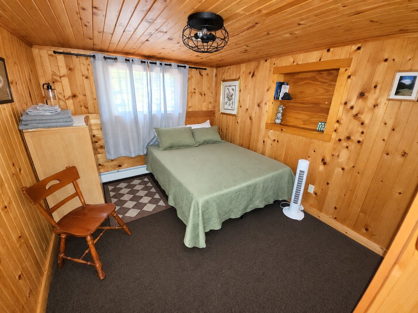 A wooden-paneled room with a bed, a chair, a radiator, and a small shelf with items.