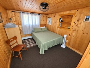A wooden-paneled room with a bed, a chair, a radiator, and a small shelf with items. - Pine Haven (Mercer)