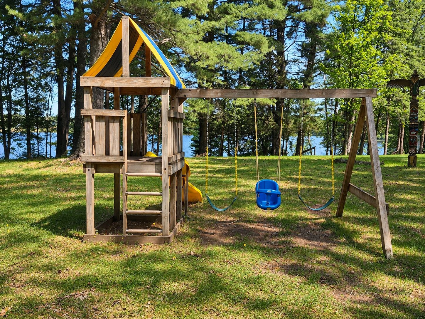 A wooden play structure with a slide and swings, set in a grassy area with trees and a body of water in the background.