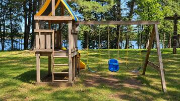 A wooden play structure with a slide and swings, set in a grassy area with trees and a body of water in the background.