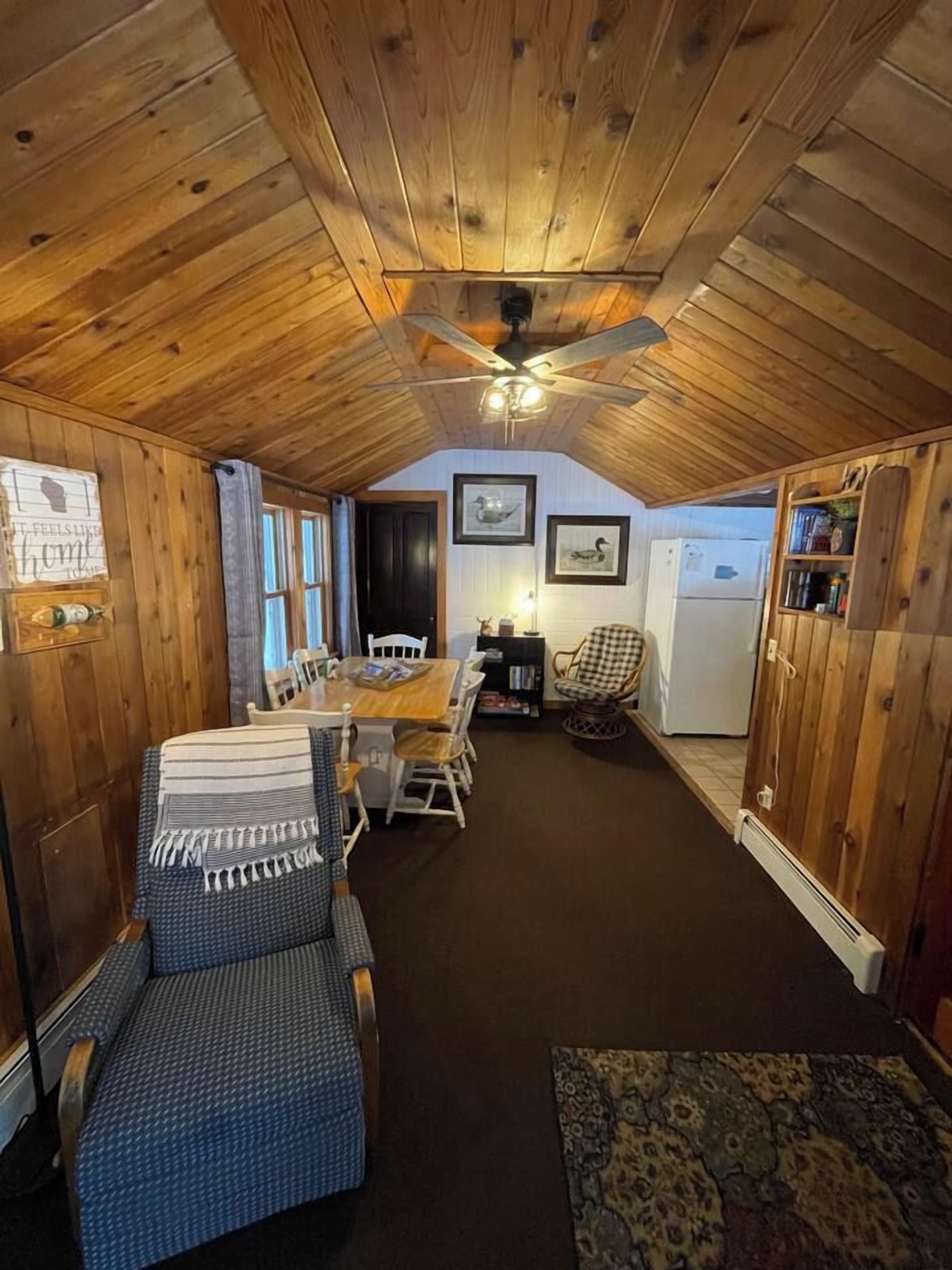 A cozy cabin interior with a wooden ceiling, a dining table with chairs, a small kitchen area, and a patterned rug.