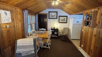 A cozy cabin interior with a wooden ceiling, a dining table with chairs, a small kitchen area, and a patterned rug.