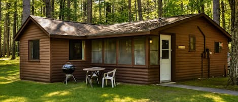 A small cabin with a porch, a grill, and a table with chairs.