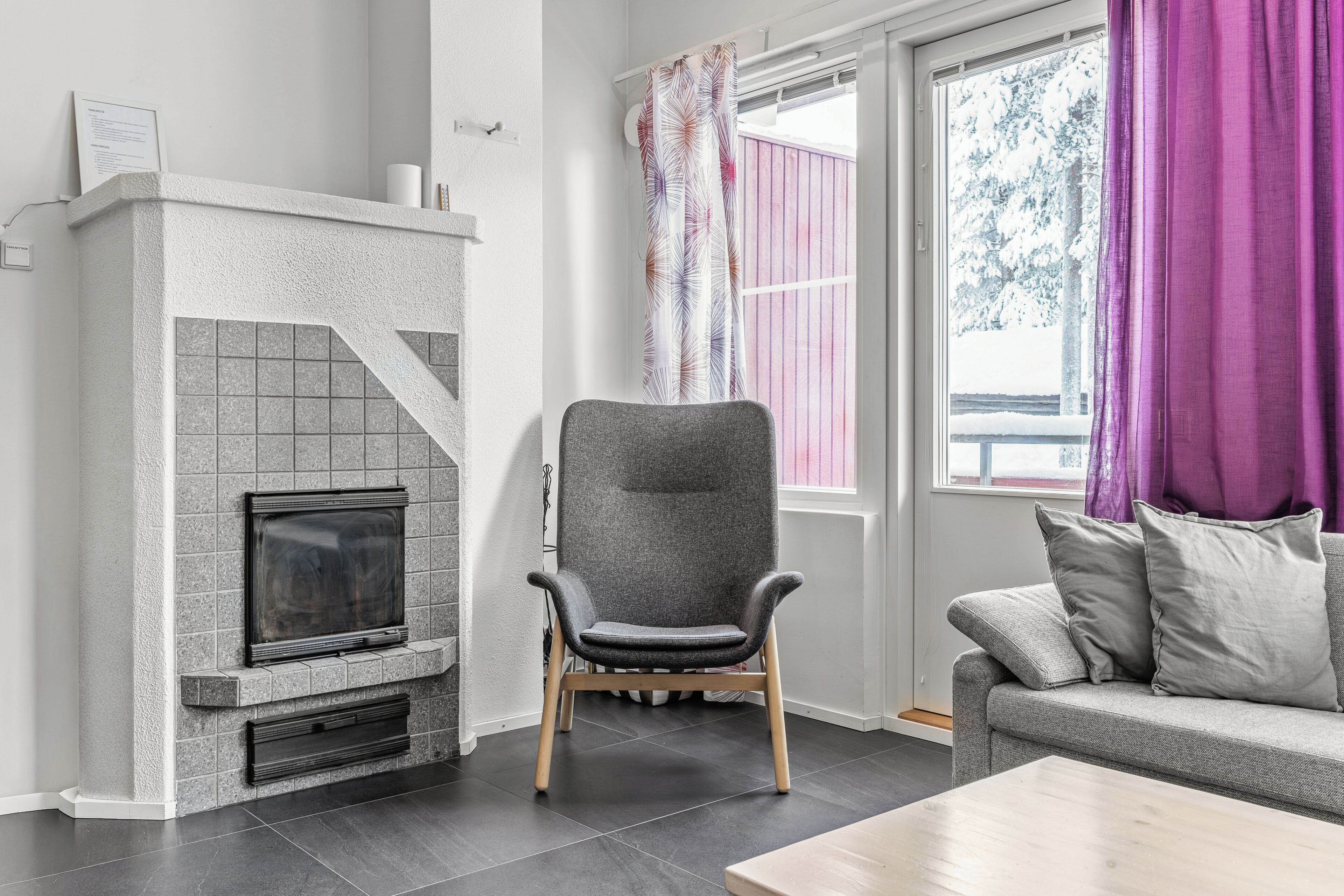 A living room with a fireplace, a grey armchair, a grey sofa, and a window with curtains.