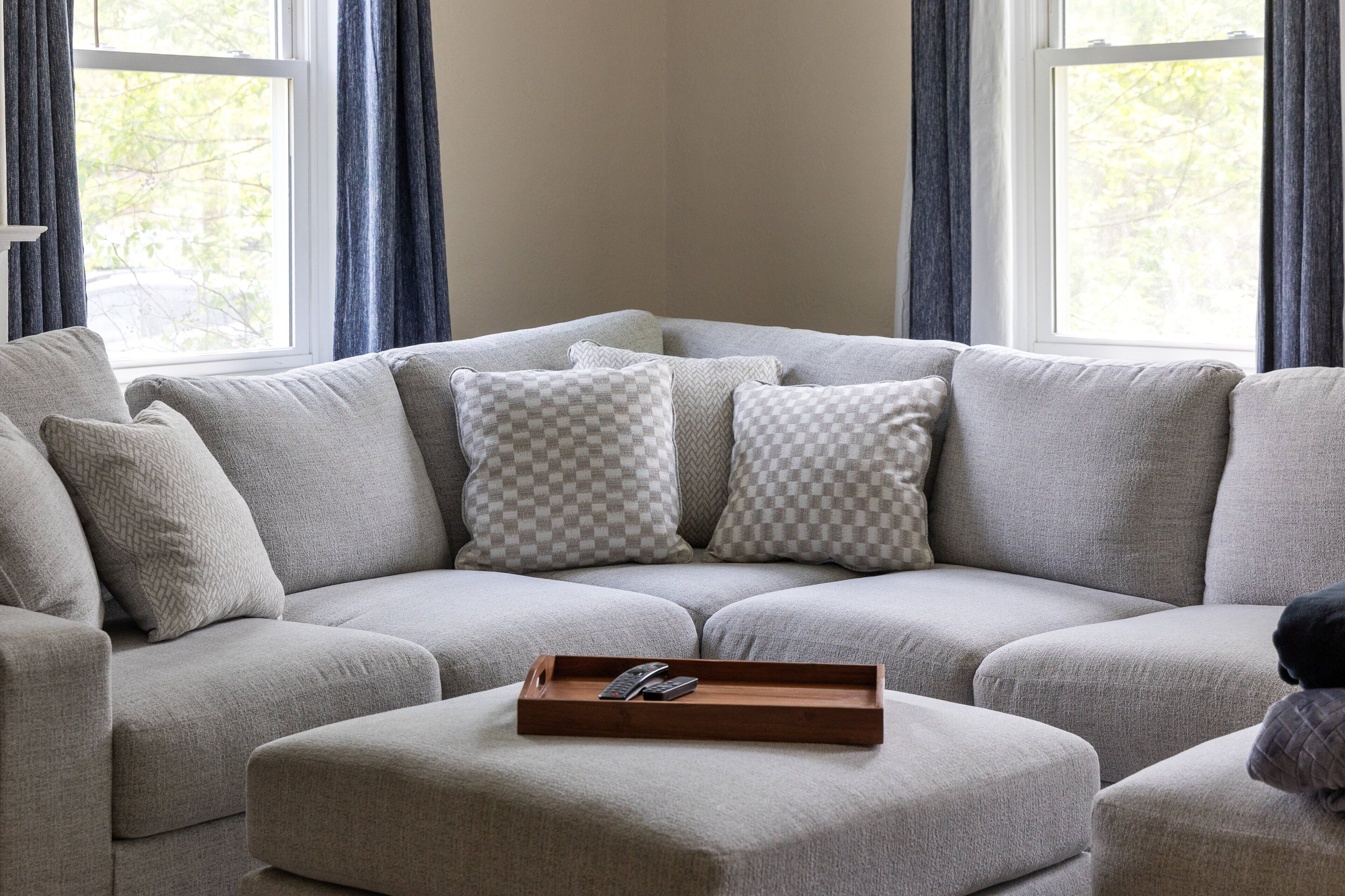 A sectional sofa with multiple pillows, a wooden tray with remote controls, and a window with curtains.