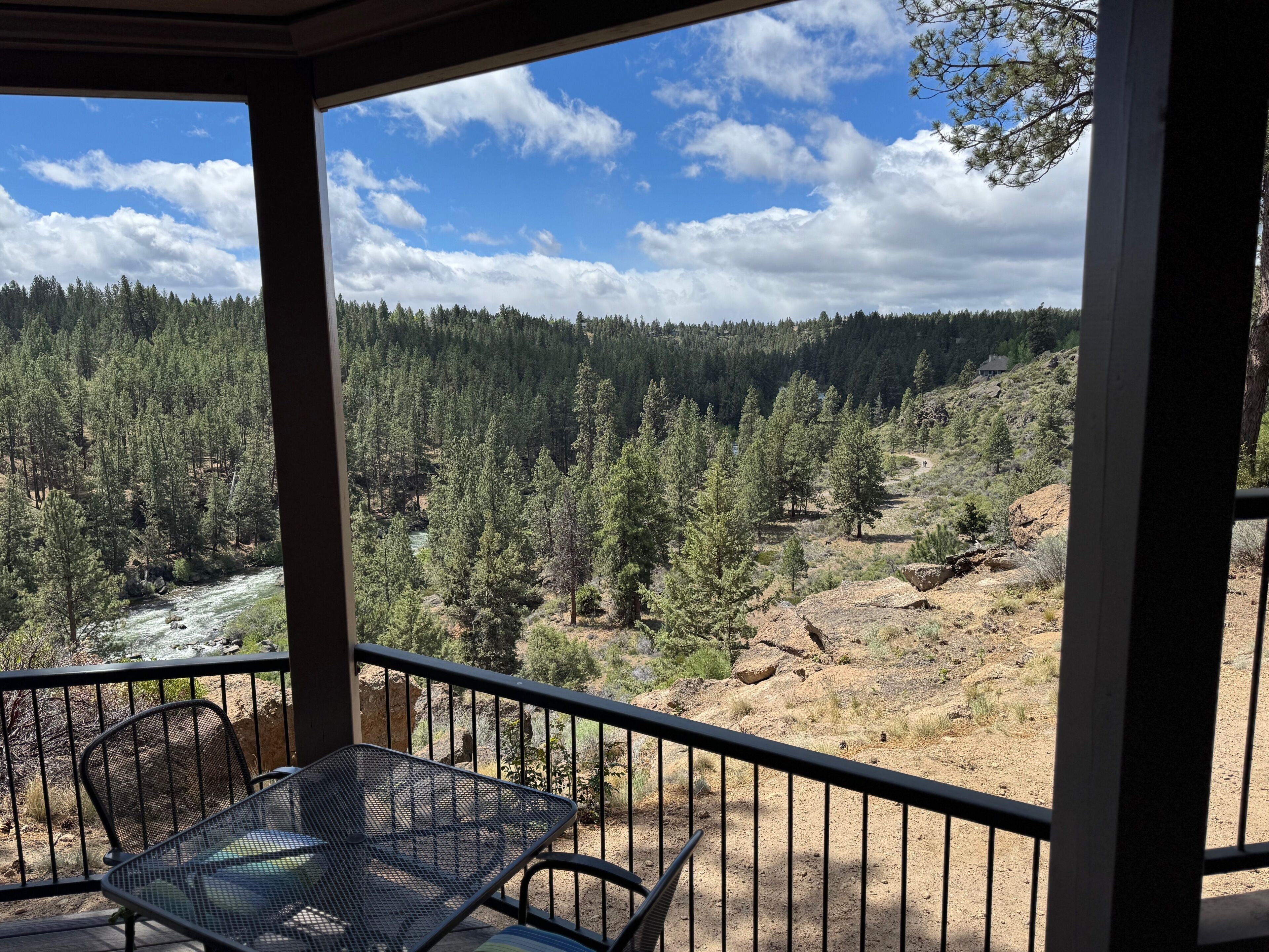 Un balcón con una mesa y sillas de metal, con vista a un valle boscoso y un río.