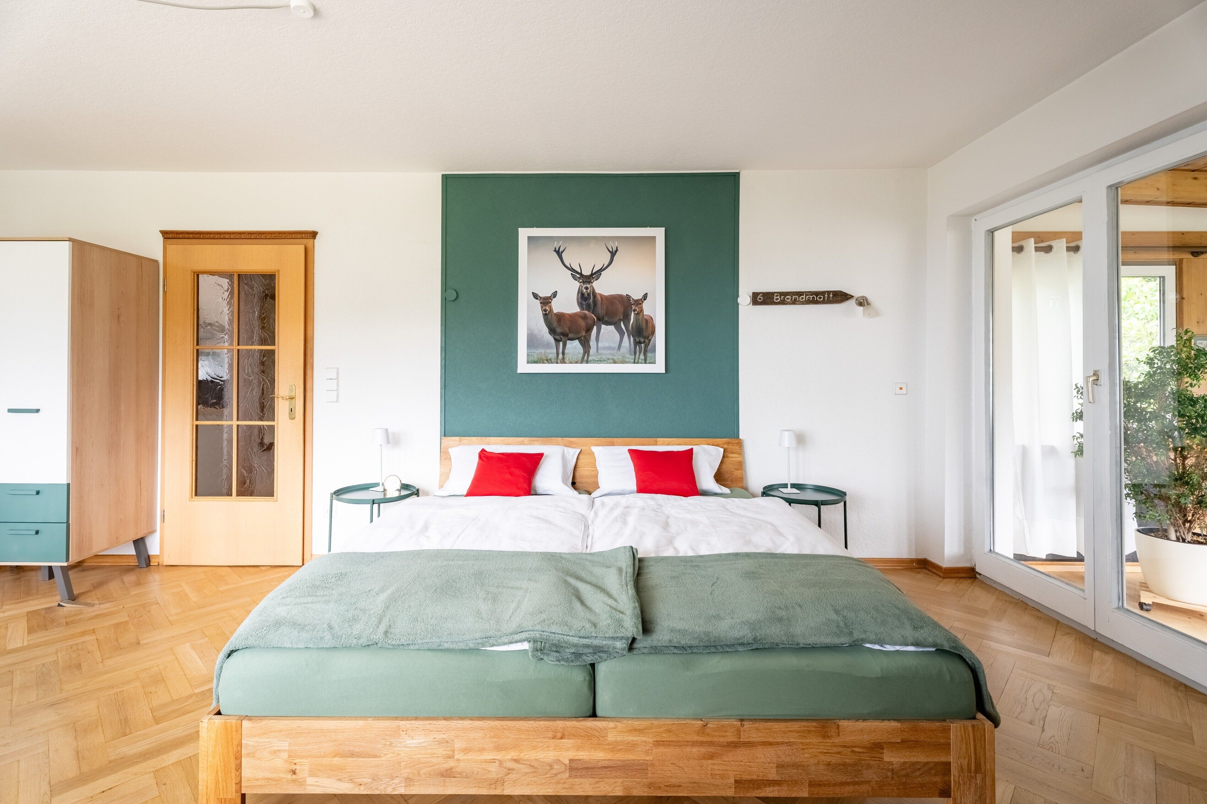 A bedroom with a bed, a wooden wardrobe, a door, a nightstand, a bedside table, a framed picture, and a window.