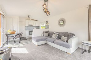 A modern living room with a sectional sofa, a dining table with chairs, a kitchen area, and a large mirror on the wall. - Host & Stay | Hazel Retreat (Felton)