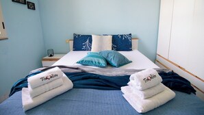 Een net opgemaakt bed met blauw-witte beddengoed, een nachtkastje met een lamp, en een muur met lijstjes.