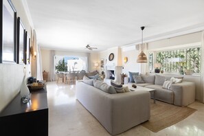 A spacious living room with a large sofa, a coffee table, and a fireplace. There is a dining area with chairs and a table visible in the background. - Elegant Villa With Pool Terrace in Marbella (Marbella)