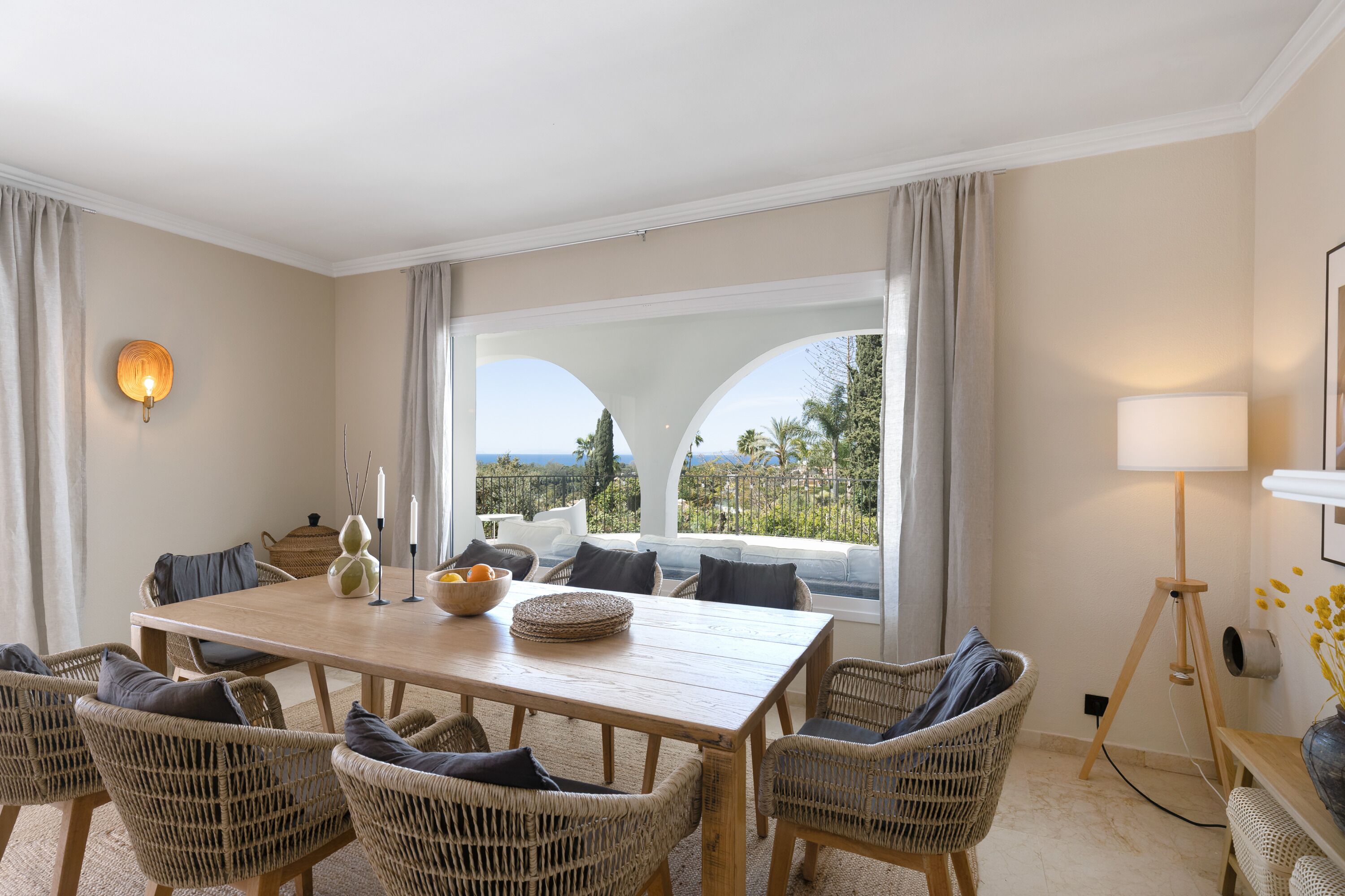 A dining area with a wooden table, wicker chairs, and a view through large windows.