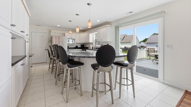 A modern kitchen with white cabinets, a central island with bar stools, pendant lighting, and a view to the outside. - Salty Paws Cottage - Where Coastal Comfort Meets Pet-Friendly Living! (Beaufort)