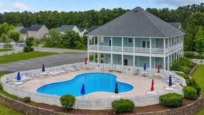 A two-story building with a swimming pool, surrounded by a white fence and featuring a covered patio area. - Salty Paws Cottage - Where Coastal Comfort Meets Pet-Friendly Living! (Beaufort)