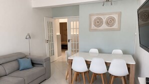 Un saló modern amb un sofà gris, una taula de dinar amb cadires blanques i un televisor de pantalla plana muntat a la paret.