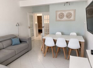 Ein modernes Wohnzimmer mit einem grauen Sofa, einem Esstisch mit weißen Stühlen und einem an der Wand befestigten Flachbildfernseher.