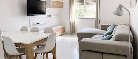A modern living room with a flat-screen TV, a sofa, a dining table with chairs, and a window with curtains.