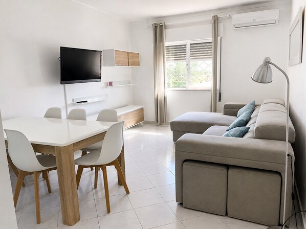 Ein modernes Wohnzimmer mit Flachbildfernseher, einer Couch, einem Esstisch mit Stühlen und einem Fenster mit Vorhängen.