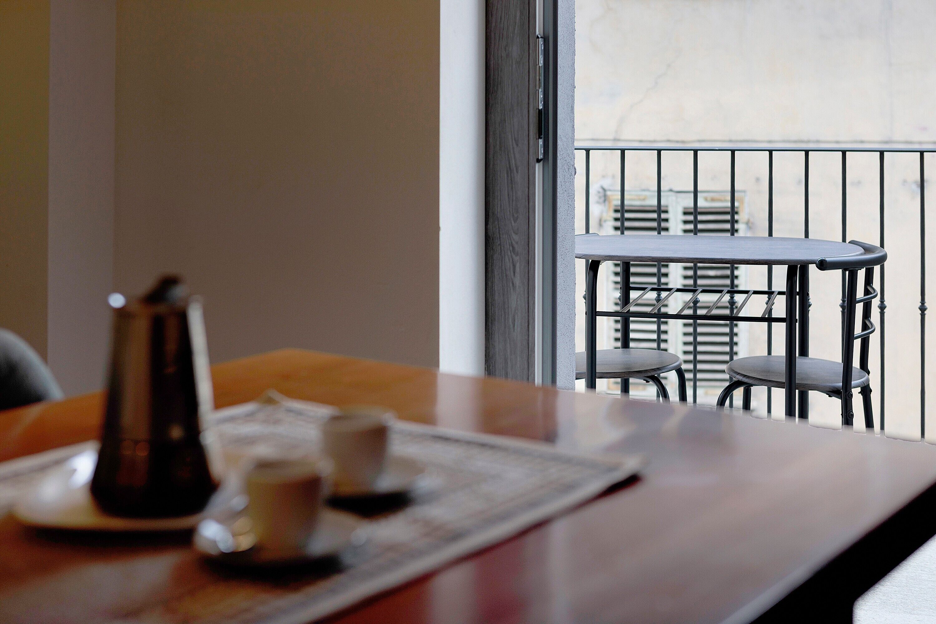 Una taula de menjador amb una cafetera, tasses i una protecció per a plats. Es veu un balcó amb barana i cadires per la finestra.