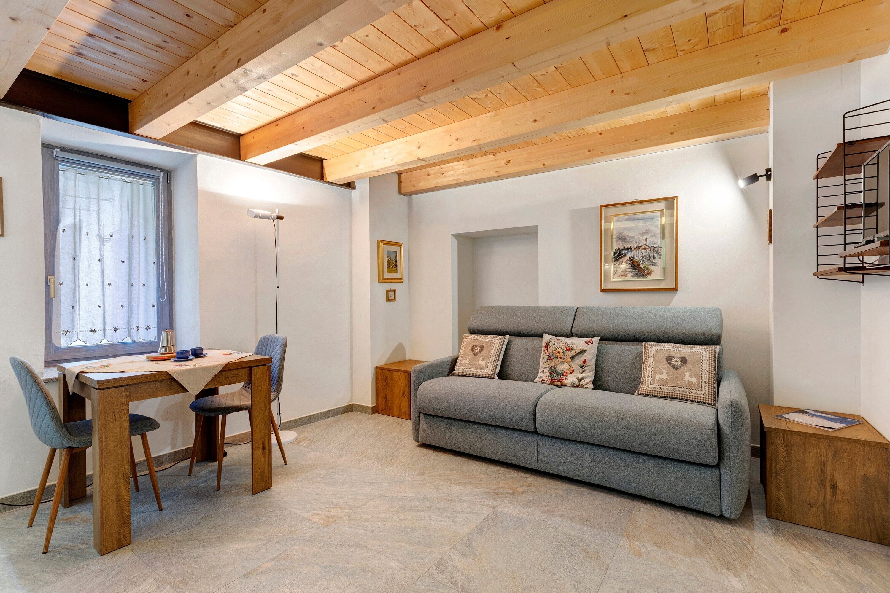 A room with a wooden ceiling, a grey sofa with decorative pillows, a wooden dining table with chairs, and a small wooden side table.