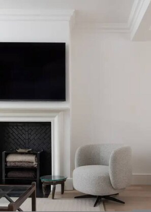 A modern living room with a fireplace, a television mounted on the wall, a grey armchair, and a small round table. - Chelsea White House Retreat 3 Bedroom House (London)