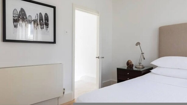 A bedroom with a bed, a bedside table with a lamp, a framed picture on the wall, and a radiator. - Chelsea White House Retreat 3 Bedroom House (London)