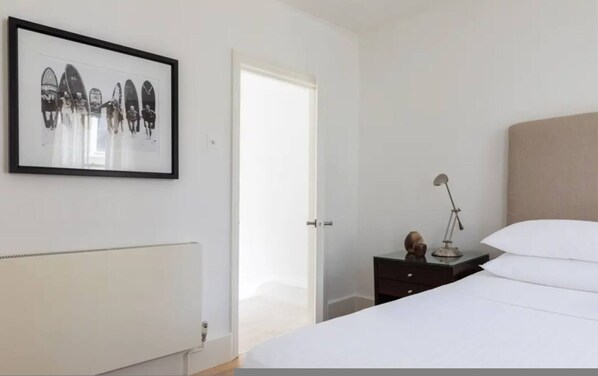 A bedroom with a bed, a bedside table with a lamp, a framed picture on the wall, and a radiator. - Chelsea White House Retreat 3 Bedroom House (London)