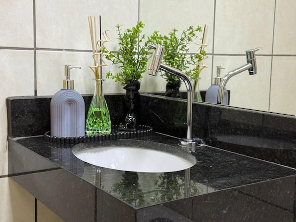A bathroom with a black countertop, a white sink, a silver faucet, a soap dispenser, and a small potted plant. - Ribeiro Flat (Várzea Grande)