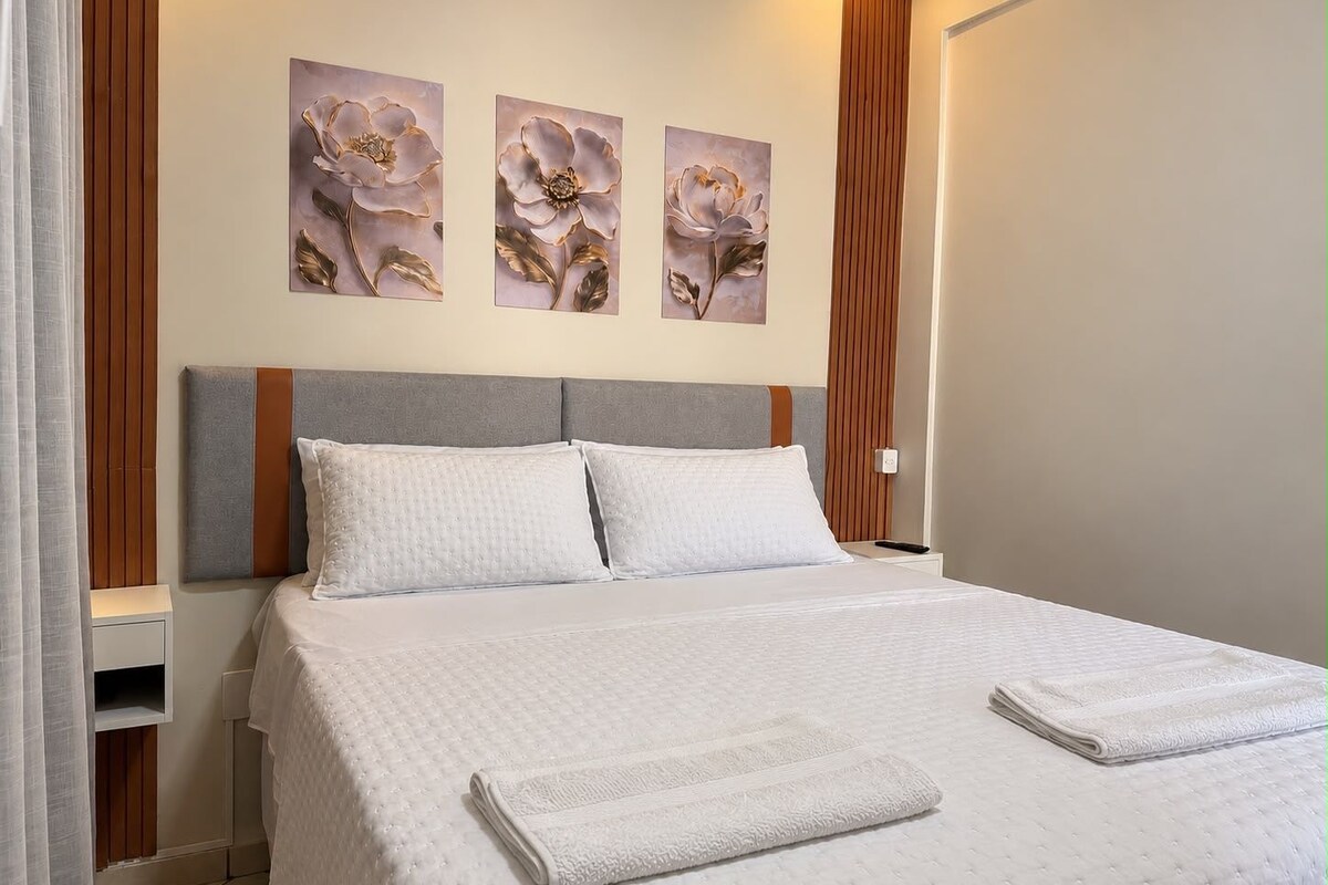 A neatly made bed with a white quilt and two pillows, a wooden headboard, and three floral paintings on the wall.