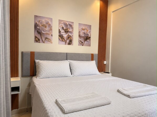 A neatly made bed with a white quilt and two pillows, a wooden headboard, and three floral paintings on the wall.