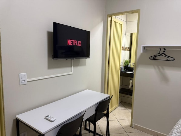 A room with a white desk, two chairs, a mounted TV displaying 'NETFLIX,' a coat hanger, and a kitchen area visible through an open door.