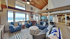 A spacious living area with a large window overlooking the ocean, a ceiling fan, and a kitchen area visible through an open door. - Akvavit | Oceanfront w/Game Room + Direct Beach Access (Ponte Vedra)