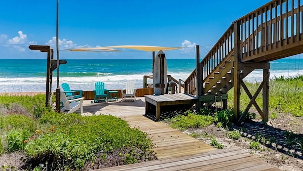 A wooden deck with chairs and a surfboard, leading to a beach with ocean views. - Akvavit | Oceanfront w/Game Room + Direct Beach Access (Ponte Vedra)