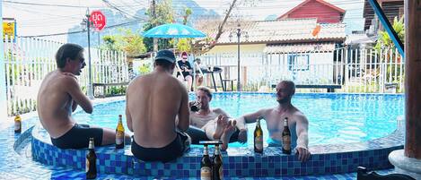 A group of people relaxing by a pool with drinks, surrounded by a tiled patio and a stop sign in the background.
