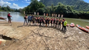 Un grupo de personas sobre una playa arenosa con kayaks y remos, un río al fondo y un paisaje montañoso.