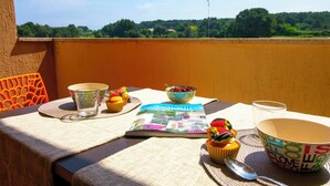 Ein Tisch mit einem Buch, einer Schale mit Kirschen, einem Cupcake und einer Tasse auf einem Balkon mit Blick auf Bäume und einen klaren Himmel.