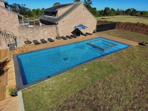 Una piscina rectangular con muelle de madera y sillones de descanso, rodeada por un edificio de ladrillo con techo inclinado.