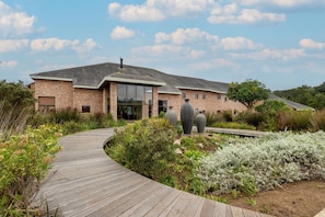 A brick building with a curved wooden pathway leading to a glass entrance, surrounded by greenery and large vases. - Plateau Country House (Nature's Valley)