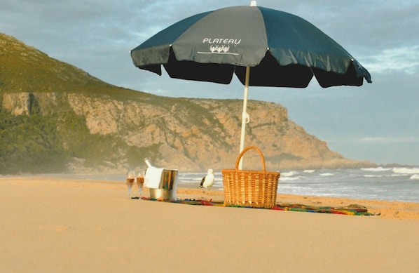 Une scène de plage avec un parasol noir portant l’inscription « PLATEAU », un panier en osier et deux seaux en métal sur le sable.