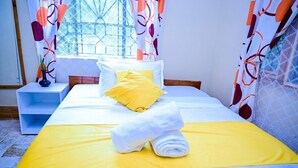 Uma cama com colcha amarela, almofadas brancas e uma almofada decorativa amarela. Há uma mesa de cabeceira branca com um vaso e uma toalha enrolada.