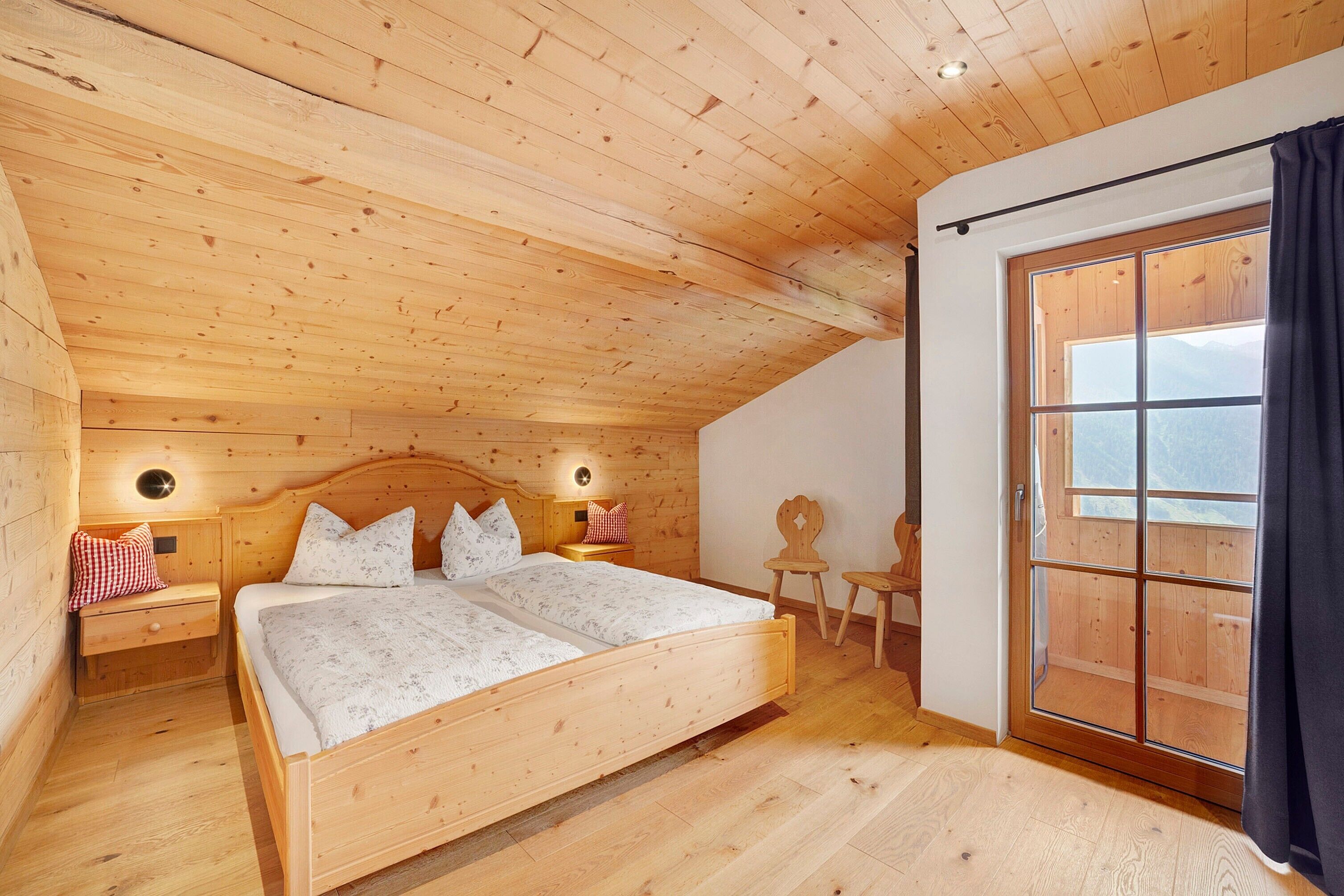 A wooden-paneled bedroom with a bed, bedside tables, a nightstand, a chair, and a large window.