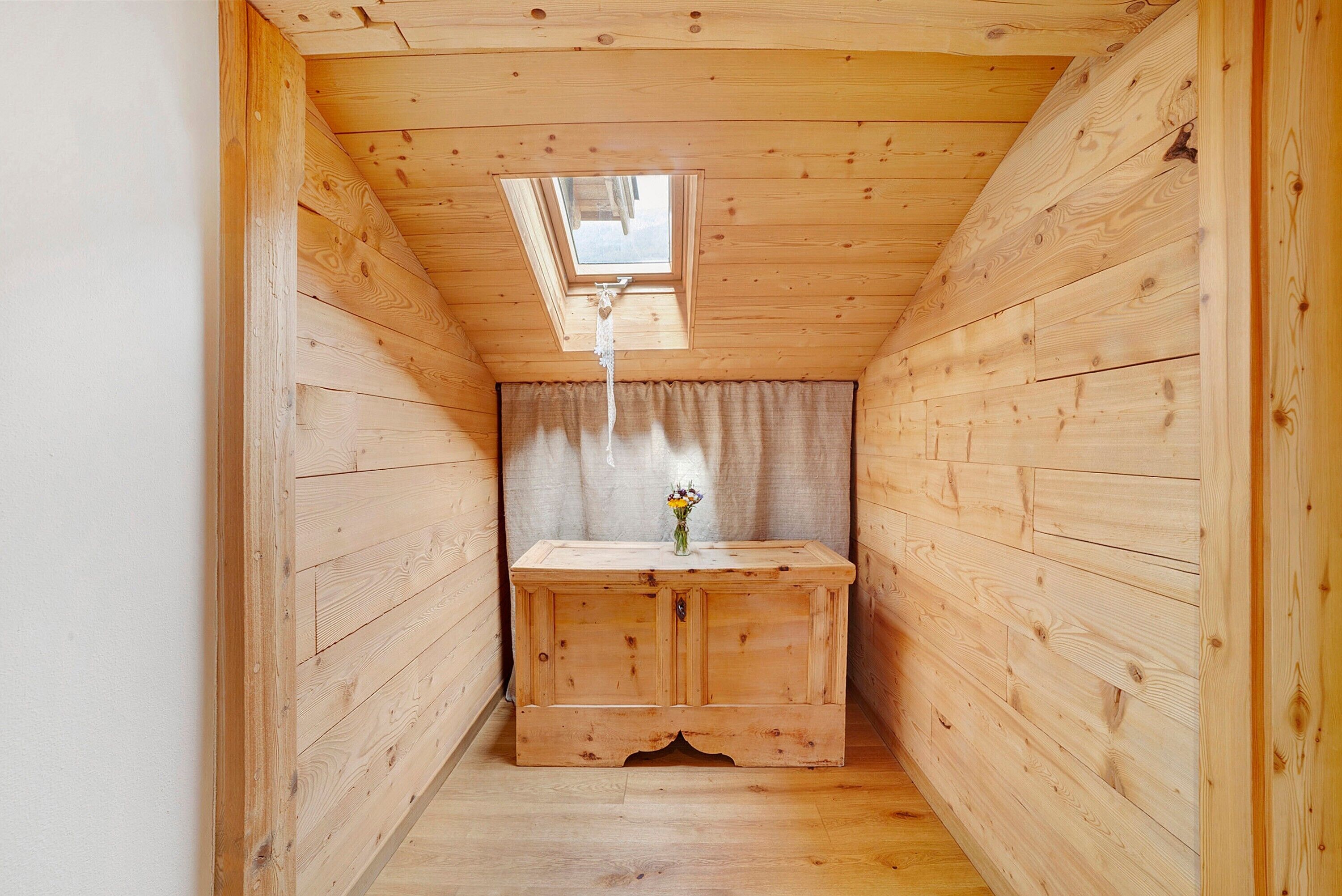 Una habitación de madera estrecha con techo inclinado, un ventanal pequeño y un armario de madera con un jarrón de flores.