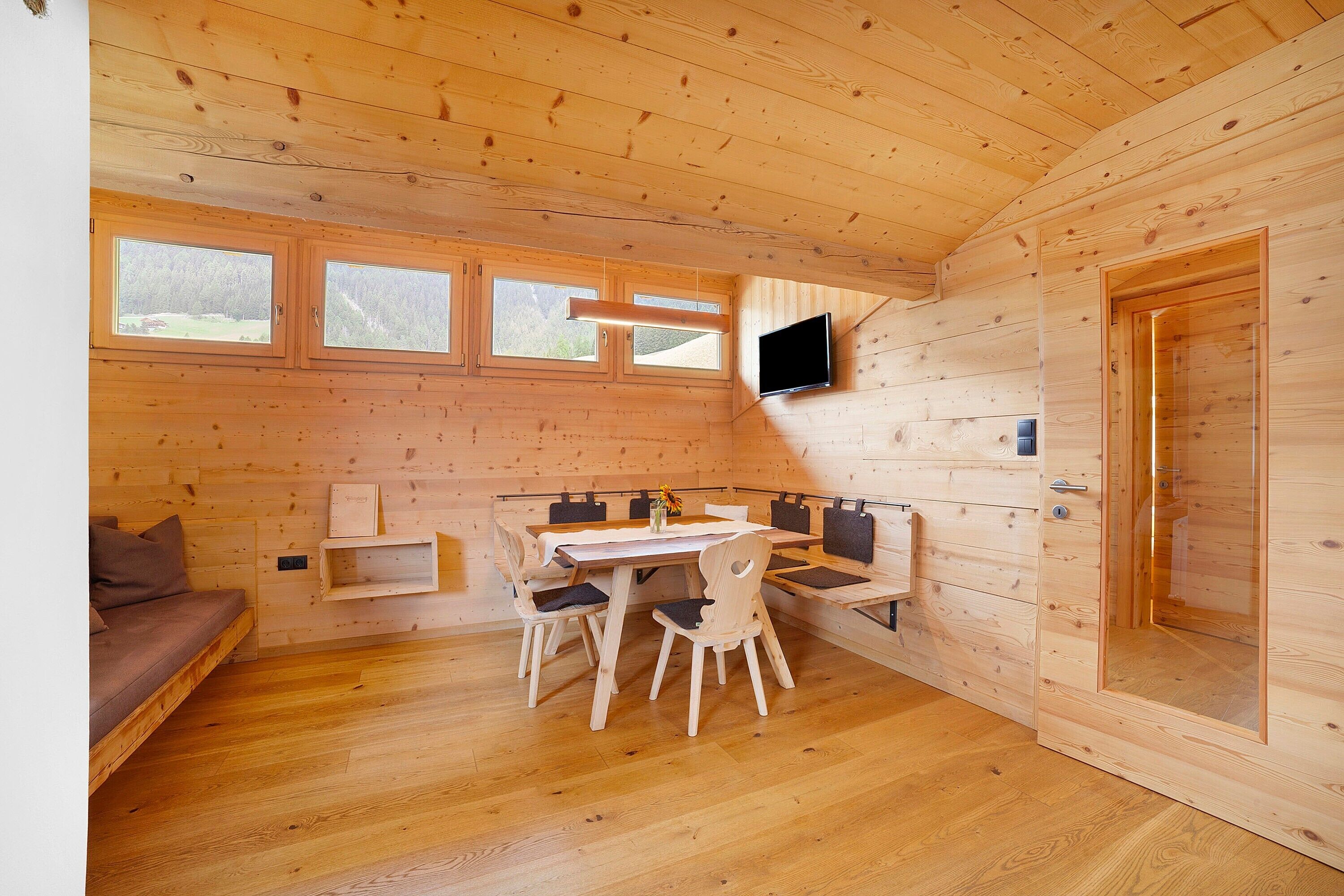A wooden cabin interior with a dining table, chairs, a sofa, a TV mounted on the wall, and a door.