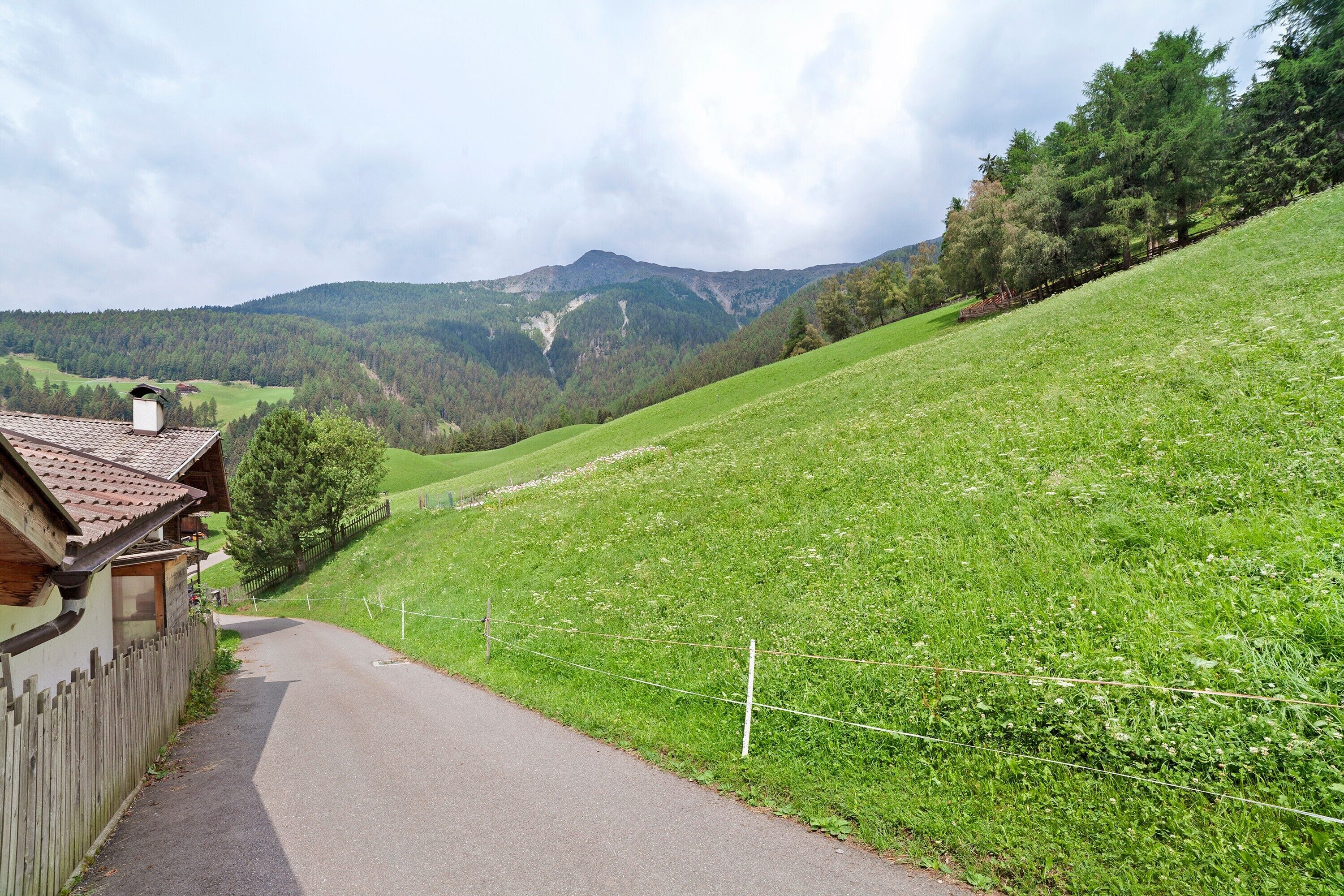 Un chemin de campagne mène à une maison en bois dotée d’un toit en pente, entourée de champs verdoyants et avec une montagne en toile de fond.