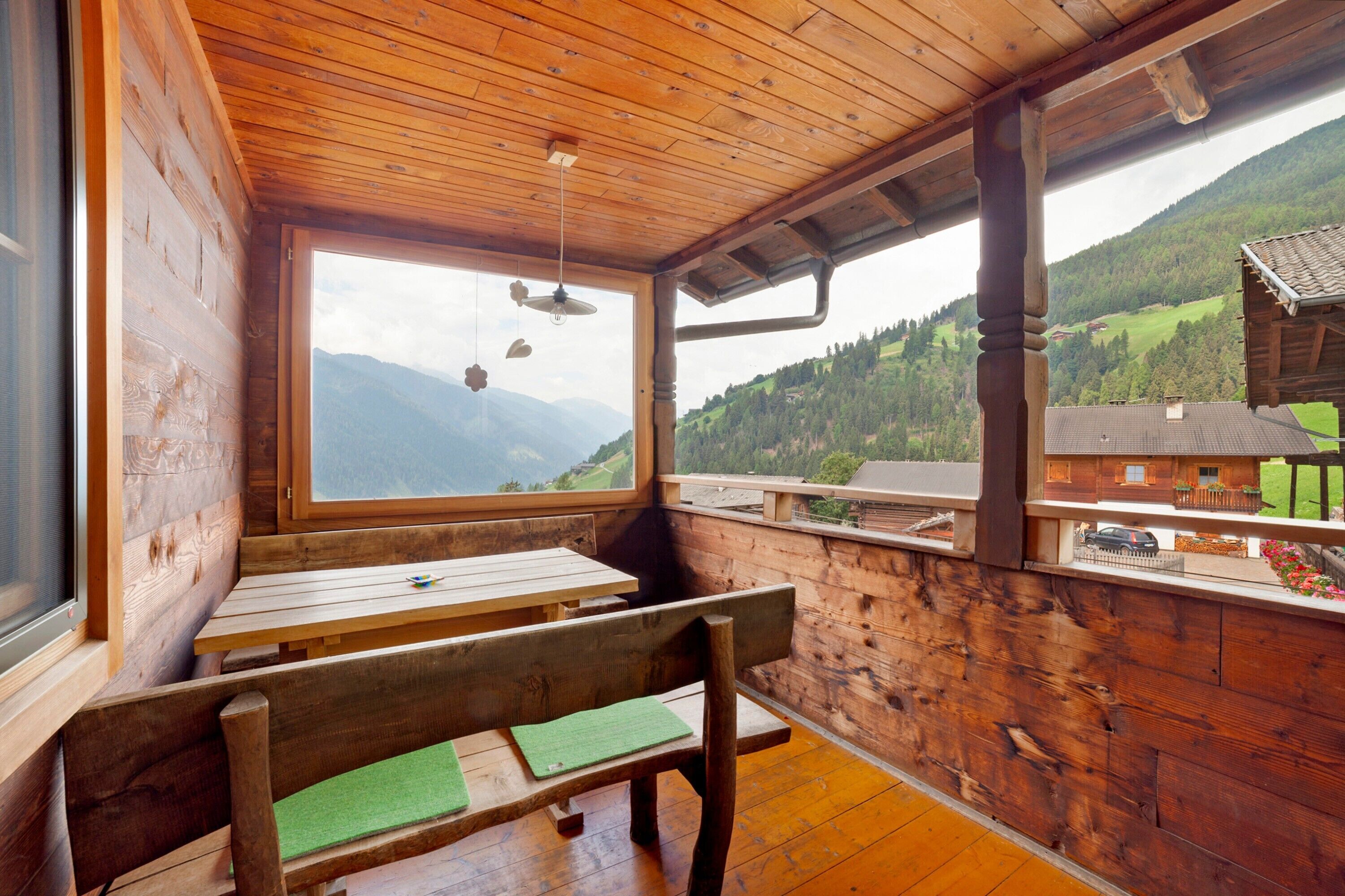 Un interior de madera con una mesa y bancos, ventanas amplias con vistas a la montaña, y un balcón con vista a un pueblo.
