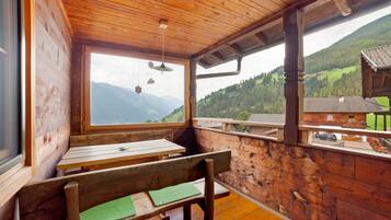Un interior de madera con una mesa y bancos, ventanas amplias con vistas a la montaña, y un balcón con vista a un pueblo.