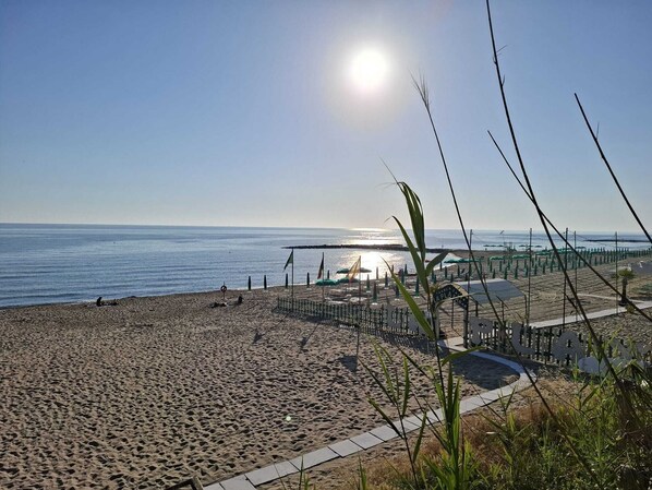 A beach with a clear sky, a sun reflecting on the water, and a fenced area with umbrellas. - Gorgeous beachfront villa, 1 hour away from Rome, parking spaces included. (Anzio)