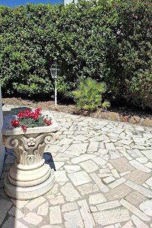A stone patio with a decorative table and red flowers, surrounded by dense green foliage and a stone wall. - Gorgeous beachfront villa, 1 hour away from Rome, parking spaces included. (Anzio)